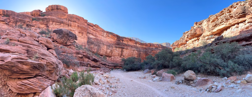 Panoramic view of of the Havasu Trail. A narrow footpath is worn through a rocky wash at the base of red rock walls.